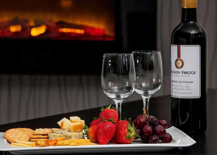 Wine bottle with platter of fresh fruit and cheese next to in-room fireplace.